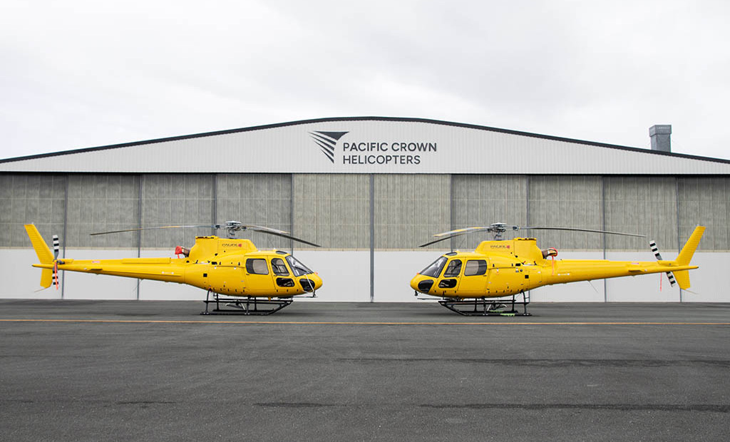 Two yellow Airbus H125 helicopters in front of Pacific Crown Helicopters hangar